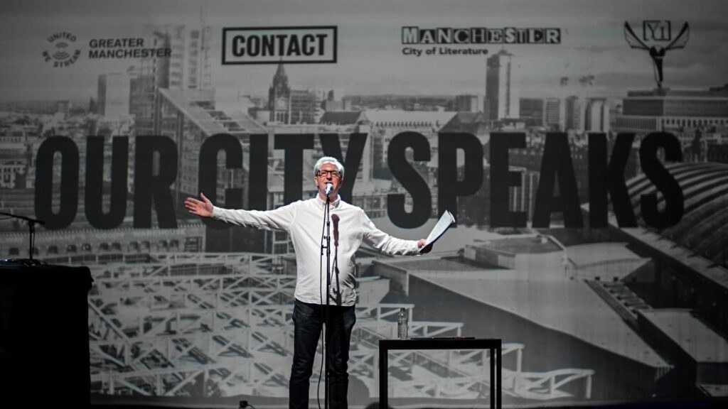 Poet Tony Walsh on stage at The Met with Our City Speaks written on the stage behind him. 